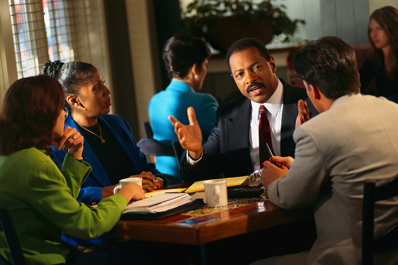 A Photograph shows a group of business people sitting round a table listening to a man, who is explaining by word and by using his hands.